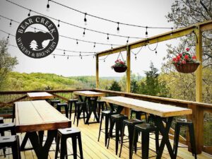 Outdoor deck at Bear Creek Lodge and Wine Co. with wooden tables, stools, string lights, hanging flowers, and scenic Ozarks views