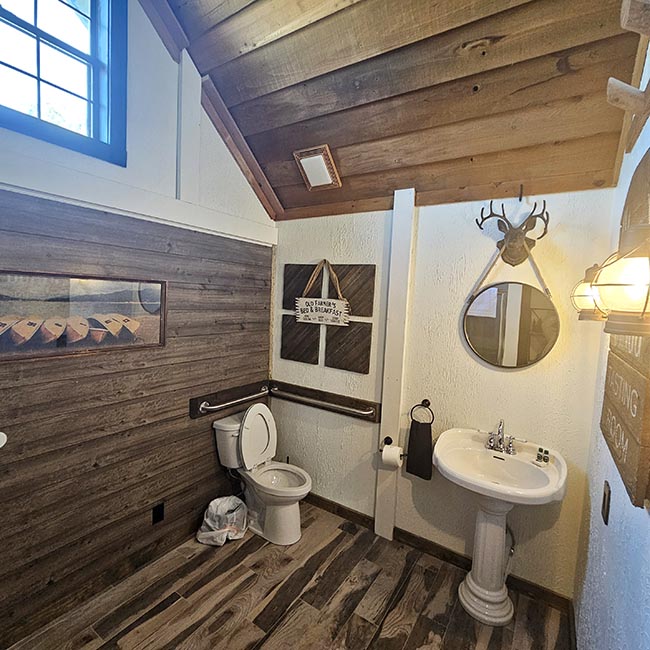 Rustic cabin bathroom at Creekside Cabin in Bear Creek Cabin Resort with pedestal sink, toilet, round mirror, wood accent walls, and a sloped wood ceiling
