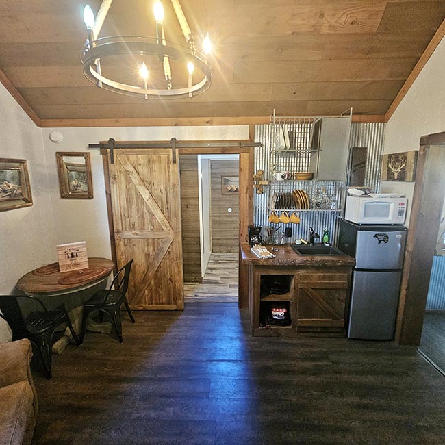 Rustic cabin kitchenette and dining area with a barn style sliding door, small dining table, mini fridge, microwave, and wood paneled vaulted ceiling at Bear...