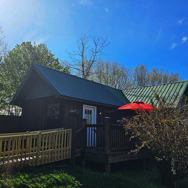 Exterior view of a Creekside Cabin at Bear Creek Cabin Resort in Walnut Shade MO, showing the front entrance, deck, ramp walkway, and a red patio umbrella un...